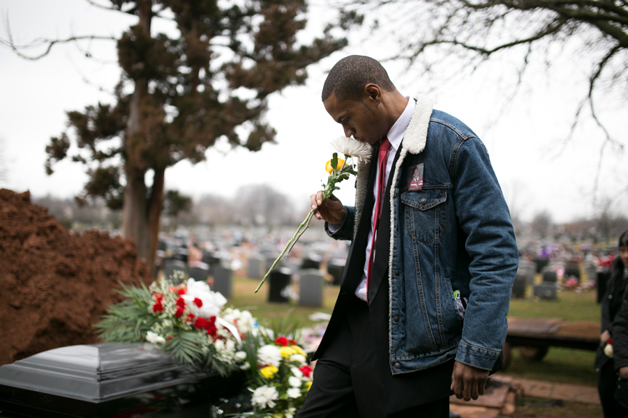 police shootings: Man in denim jacket over dark suit sniffs flower next to casket covered with flowers.