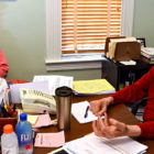Florida: 2 men with glasses sit at desk in office; one at left, with gray beard and mustache, wears long-sleeved orange T-shirt, flag-patterned hat; the one at right is bald, wears long-sleeved red T-shirt.