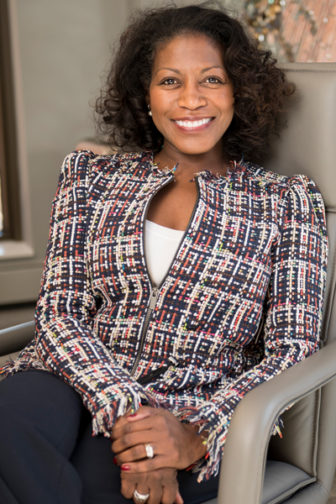Smiling woman with curly dark hair wearing zippered print jacket sitting in executive office chair.