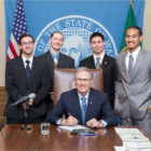 SAGs: 4 smiling young men stand behind desk chair that smiling man sitting in at desk holding pen. U.S., state flag in background.