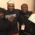 tips: Man with dark beard, mustache, holding certificate and wearing glasses, hat and dark blue Georgetown University sweatshirt is hugged by elderly couple on either side of him.