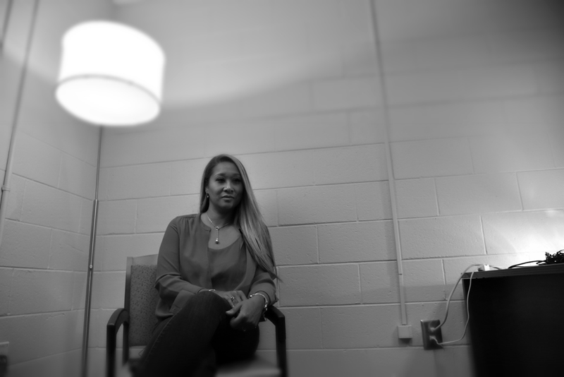 homelessness: Woman with long hair, necklace sits under lamp against cinderblock wall; black and white photo