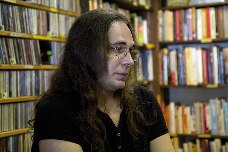 trans: Person with long brown hair, glasses, short-sleeved black top looks thoughtful sitting in bookstore