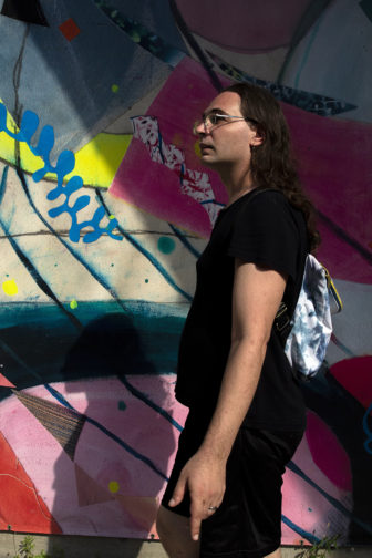 trans: Person with glasses, long brown hair, backpack walks past mural