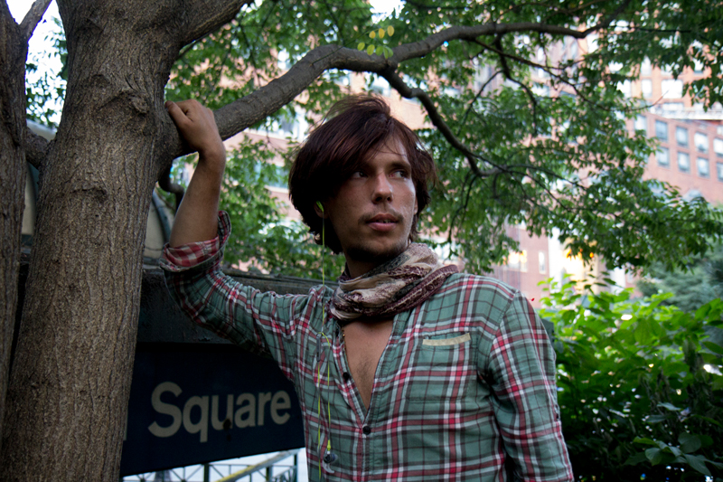 LGBTQ: Man in plaid flannel shirt, scarf looks to right holding on to tree.