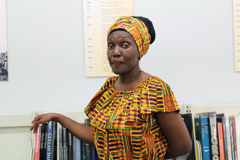 fines: Woman in matching colorful dress, head wrap, rests right arm on books on shelves.