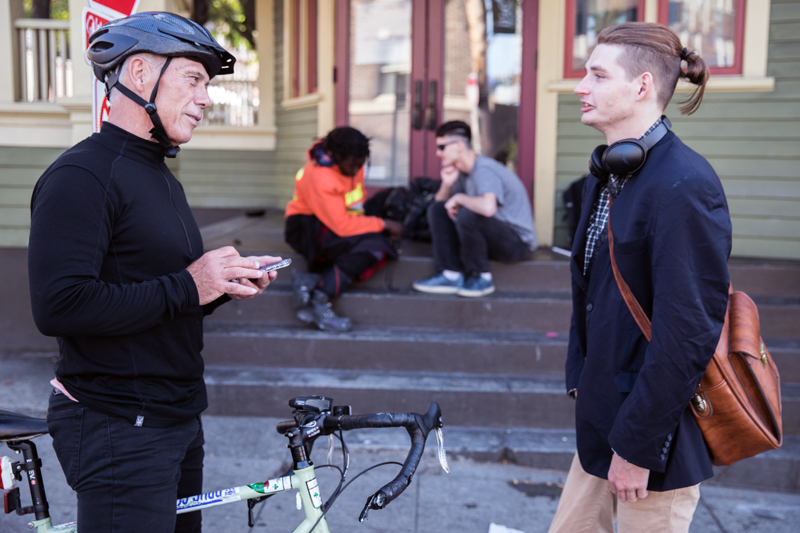homelessness: Man in black with bike helmet on talks to man with headphones around neck, messenger bag on back.