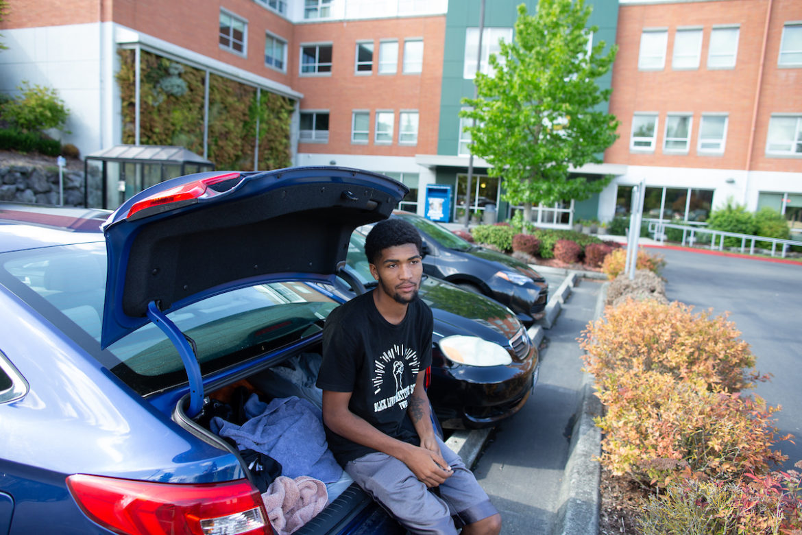 Tacoma: Depressed-looking young man with dark blue T-shirt, light blue shorts sits on edge of car trunk that has towels inside.