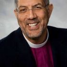 bishop: smiling man with short dark hair, scruffy beard, mustache, wearing clerical collar with maroon robe, dark jacket