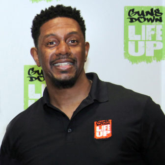 Guns Down Life Up: Smiling man with black polo shirt with orange patch that says life up stands in front of white wall with logos that say guns down life up