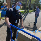 Man in dark blue uniform, mask, holds cart full of boxes. Woman in blue T-shirt, shorts in background.