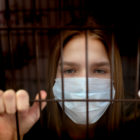 collaborate: Sad teenager girl in medical mask behind bars.