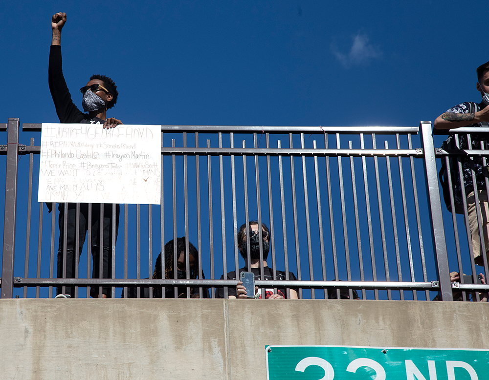 Philadelphia: Person wearing mask, sunglasses raises fist from overpass, holds sign.