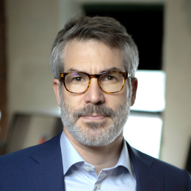 Serious-looking man with graying hair, mustache, beard, wearing glasses, dark blue jacket, light blue shirt.