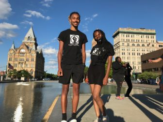 Syracuse: Young man and woman wearing matching black T-shirts, shorts, smile and pose on sidewalk