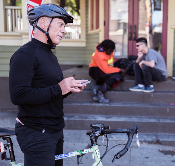 homeless shelter: Man in black sweatshirt and pants wearing black bike helmet stands in front of building steps holding bike looking down street