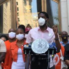 gun violence: Man in short-sleeved pink shirt, mask speaks at outdoor podium surrounded by crowd wearing masks