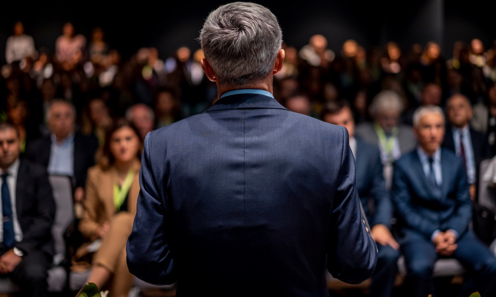 Collaboration: Man in suit making a speech in front of a big audience