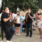Schenectady: Man in black with megaphone wearing mask talks to small group wearing masks, holding up phones. One woman is wearing superhero-style cape.