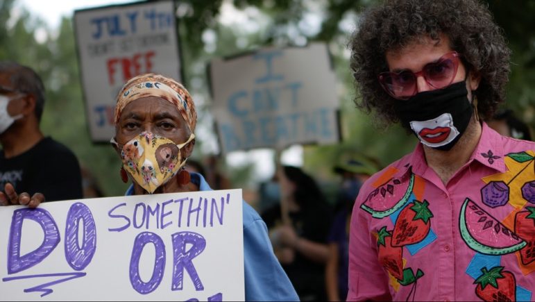defunding: Man and woman wearing colorful clothes, masks, woman holds up partly obscured sign that says do somethin’ or
