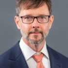 Gary Green headshot wearing dark-framed glasses, nvy suit jacket with whit shirt and coarl tie