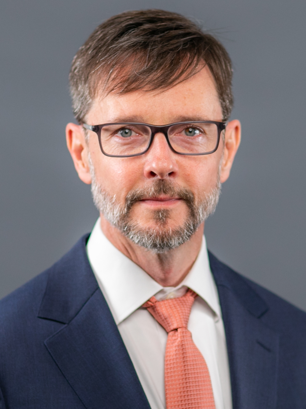 Gary Green headshot wearing dark-framed glasses, nvy suit jacket with whit shirt and coarl tie