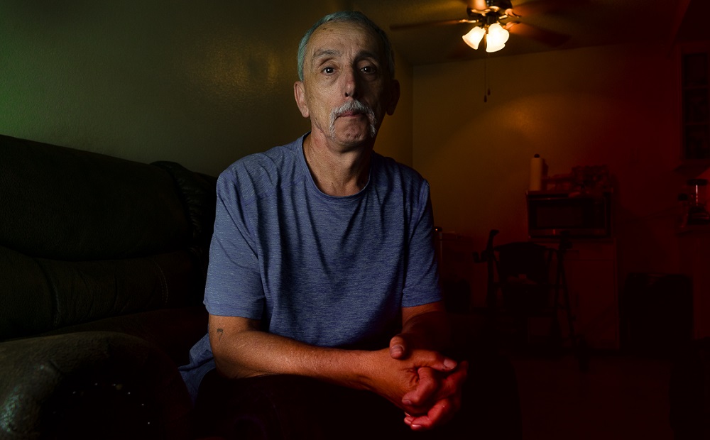 Gladiator School: Man with short gray hair, long droopy mustache, blue T-shirt sits in dark room.