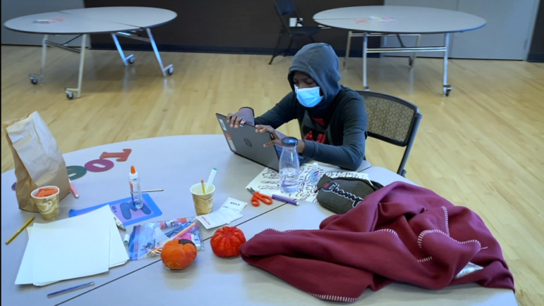 homeless: Student wearing mask, hoodie sits at round table covered with laptop, school supplies