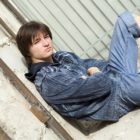Teenage boy wearing jeans rolled to knee, jeans jacket sitting on ground outside alone, looking wary