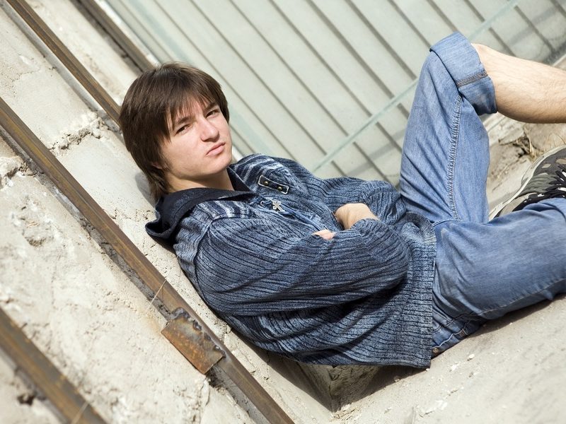 Teenage boy wearing jeans rolled to knee, jeans jacket sitting on ground outside alone, looking wary