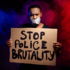 police brutality: Man of color holds sign that says stop police brutality. Adhesive tape covers his lips. Red and blue smoke in background