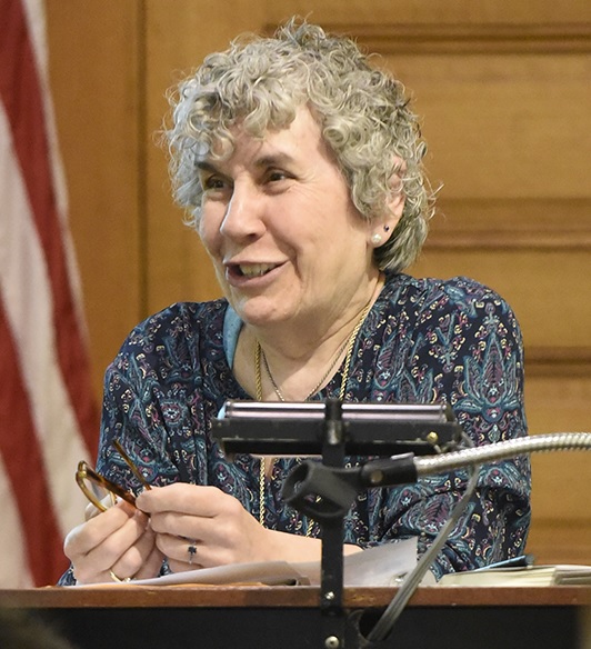 book club: (headshot), Jane Guttman, retired correctional educator, smiling woman with short, curly gray hair at lectern