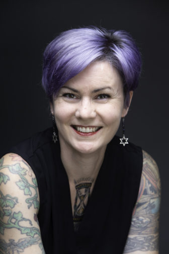 white nationalism: Nora Flanagan headshot - smiling woman with lilac hair, full sleeve tattoos wearing black tank top, earrings
