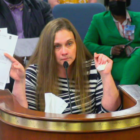 South Carolina juvenile detention reform: Woman with long light brown hair wearing black and white striped top holding papers up with her right hand stands behind a wood and brass podium speaking into a microphone.