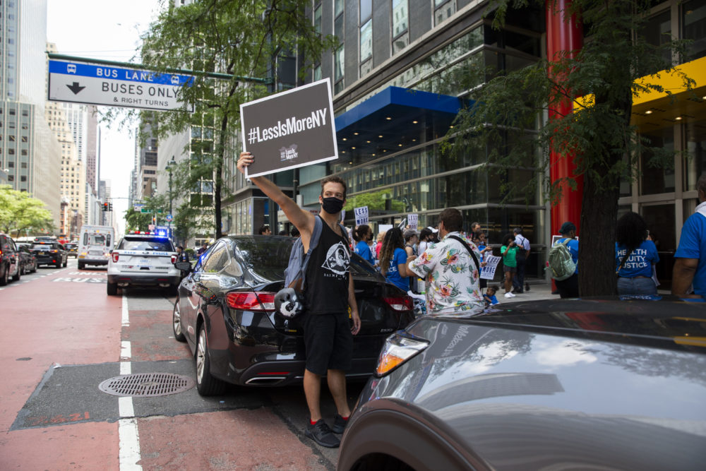 #LessIsMore Rally NY: Man in black shorts and tank top holding #LessIsMoreNY sign stands in bus lane of busy NY street in front of crowd with similar protest signsstanding on sidewalk