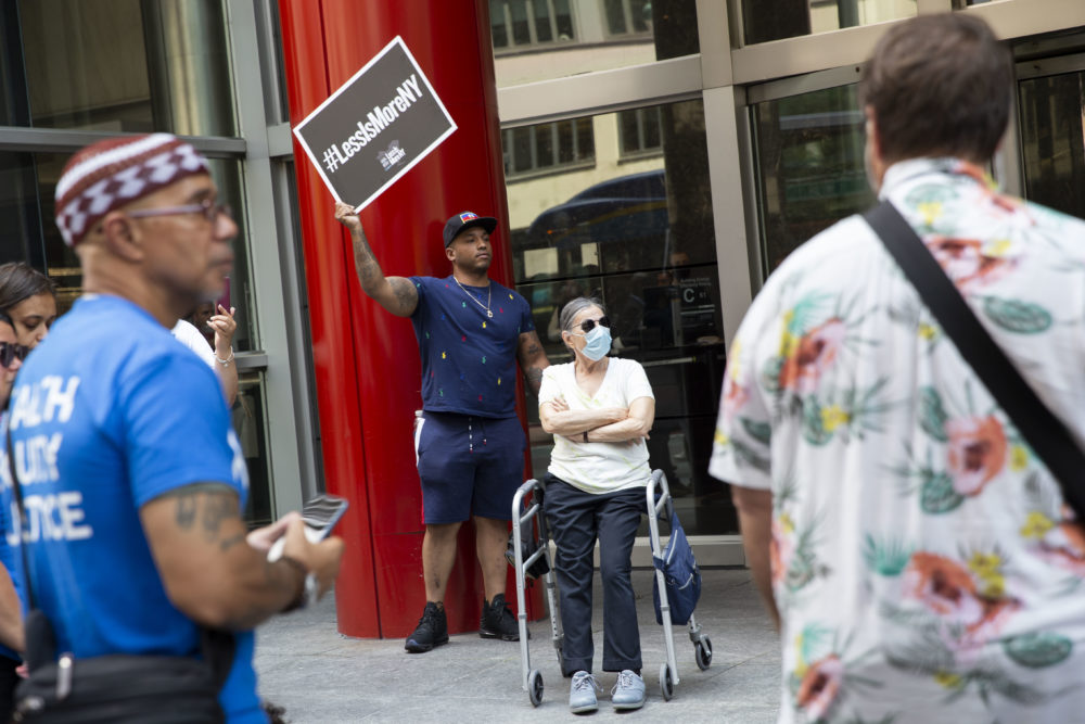 #LessIsMore Rally NY: Manin navy shorts and t-shirt with #LessIsMoreNY sign stand on sidewalk in front of red pillar of building windows next to a woman sitting in walker