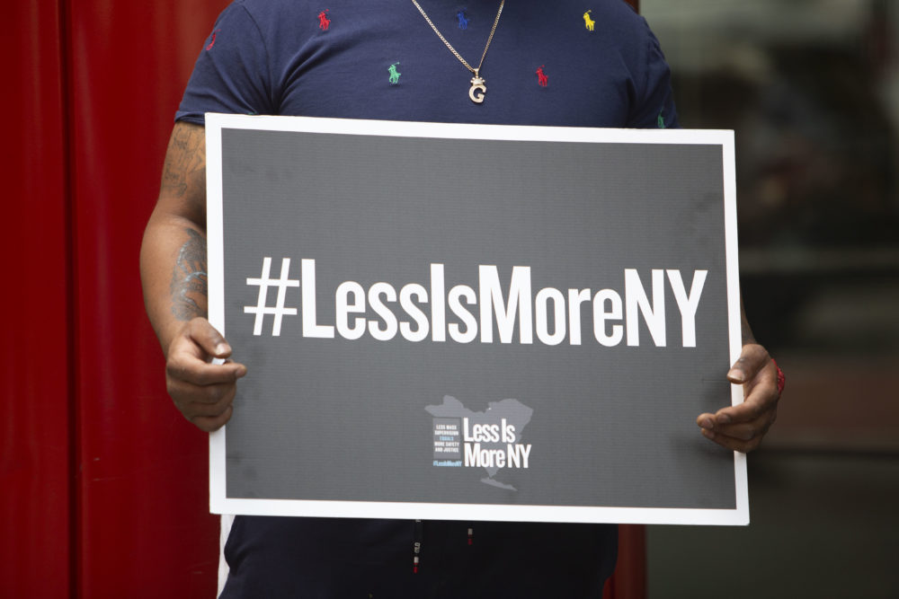 #LessIsMore Rally NY: Closeup of sign #LessIsMoreNY sign with white text on charcoal background held my man in navy t-shirt standing in front of red pole