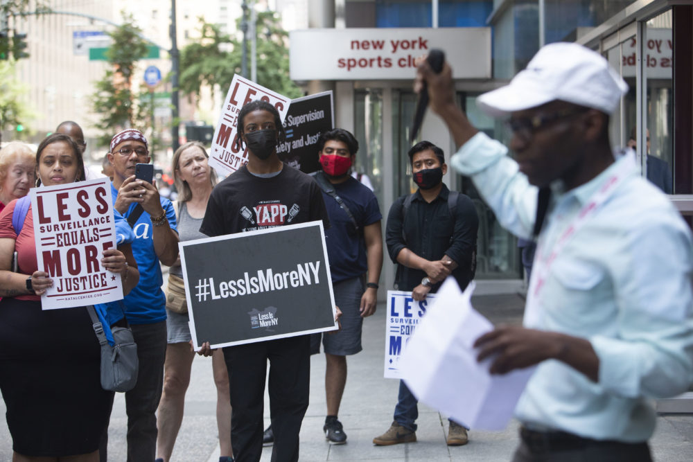 #LessIsMore Rally NY: Crowd with Less Is More signs stand on sidewalk listning to Black man in white shirt and baseball cao holding microphone