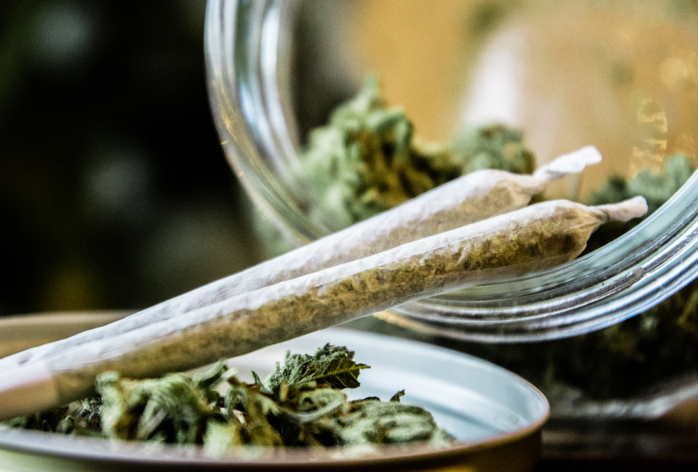 NY marijuana laws: Two hand-rolled marijuana joints rest on lid and glass jar lying on its side with marijuana buds in it.