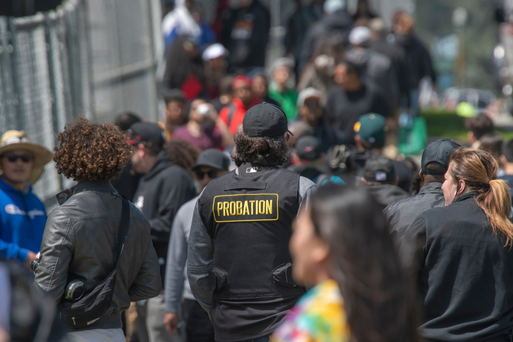 Juvenile diversion: Person wearing black baseball cap and black vest with patch reading PROBATION in yellow letters on backrowd of people walking on sidewalk