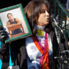 Gun shooting statistics: Woman speaking into microphones with dark hair wearing white top with black jacket and orange and purple long ribbons around her neck stands holding a green-framed photo of young man