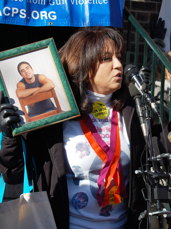 Gun shooting statistics: Woman speaking into microphones with dark hair wearing white top with black jacket and orange and purple long ribbons around her neck stands holding a green-framed photo of young man