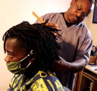Savannah gun violence: Black man in gray shirt gives haircut to a young Black man with dreadlocks wearing mask 