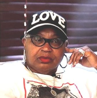 Savannah gun violence: Black woman in baseballcap with LOVE in rhinestones in white t-shirt sits with head leaning against hand