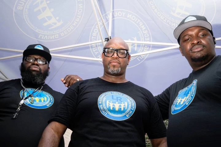 Harlem Youth Hub: Three bBlack men wearing navy t-shirts with same graphic on front stand looking into camera in front of light blue background.