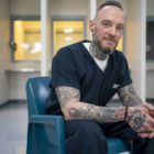 New Mexico jail: Michael Brown portrait with short light brown hair and beard smiles into camera sitting in turquoise plastic chair in prisoner visiting room wearing navy blue prison uniform with heavily black tatooed arms on lap and fingers interlaced.