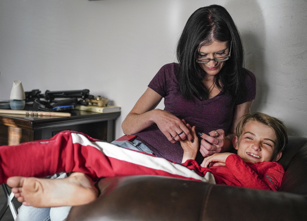 New Mexico jail: Woman with long dark hair wearing dart top and glasses sits on couch tickling pre-teen blonde boy wearing red long-sleeved top with red and white sweatpants