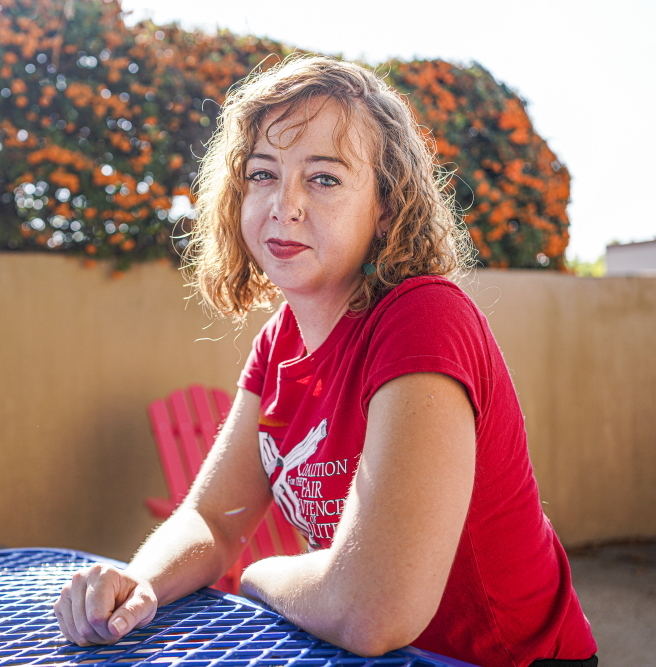 New Mexico jail: Denali Wilson headshot of curly-haired blonde woman wearing red t-shirt sitting at blue table in yard with tan adobe wall in front of orange blossoming shrubs