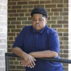 Police excessive force children: Young Black teen boy wearing blue polo shirt stands in front of red brick building leaning on metal sidewalk railings