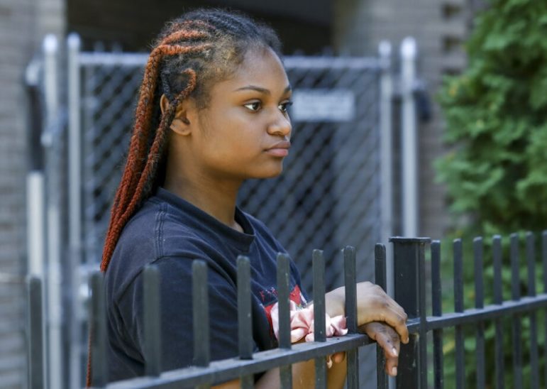 Police excessive force children: Black teen girl wearing black t-shirt with long red braids stands staring into the distance over top of black metal fence